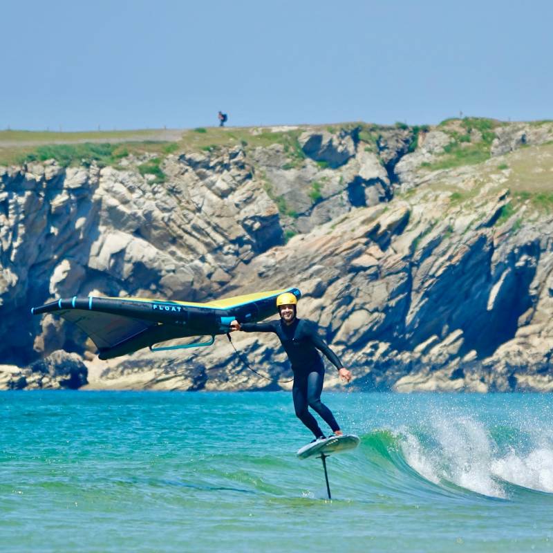 Personne en combinaison et casque jaune pratiquant le wingfoil sur une planche à foil, volant au-dessus de l'eau turquoise devant des falaises rocheuses