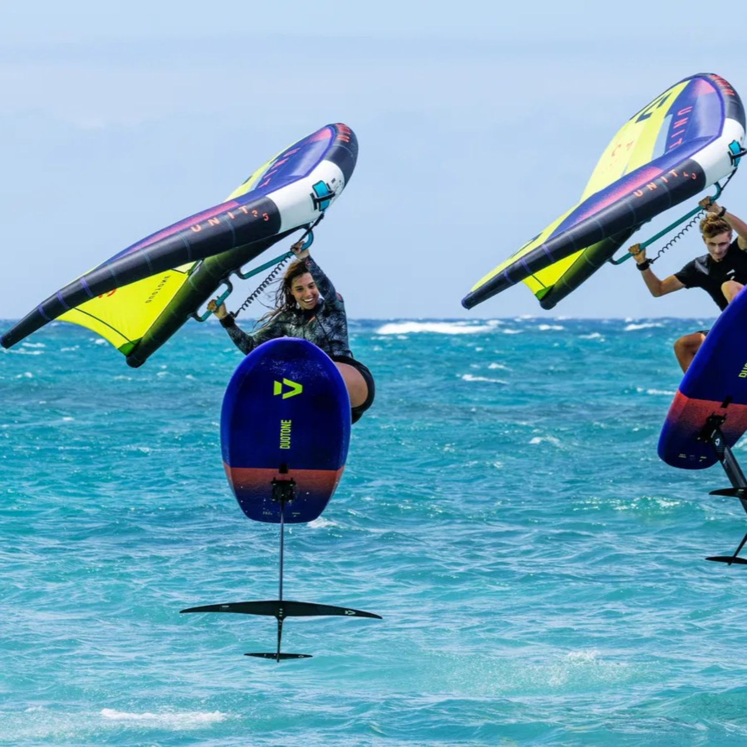 Deux riders en wingfoil utilisant des ailes Duotone Unit avec poignées Handle Fusion Set 2025, sur foil au-dessus de l’eau turquoise