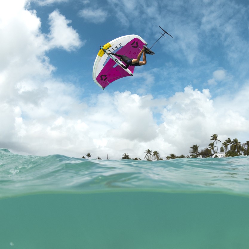 Rider en plein vol sous un ciel ensoleillé après un saut, suspendu au-dessus de l’eau avec la wingfoil Duotone Unit SLS 2025 coloris blanc et rose.
