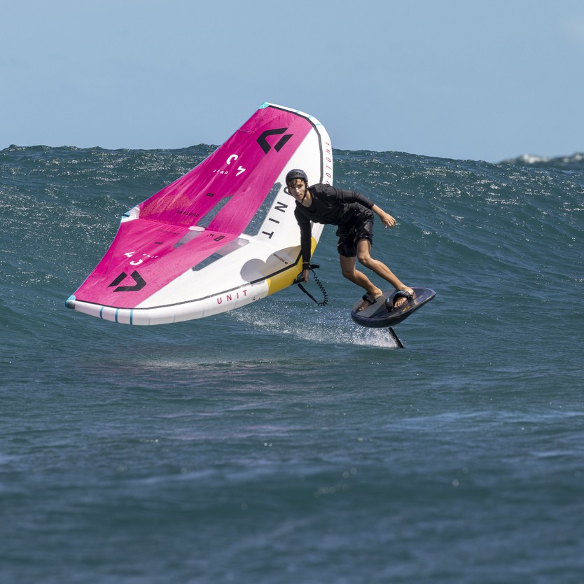 Rider en action sur une mer à vagues modérées, maniant une wingfoil Duotone Unit SLS 2025 au nouveau coloris rose et blanc, avec des éclaboussures d’eau autour de la planche