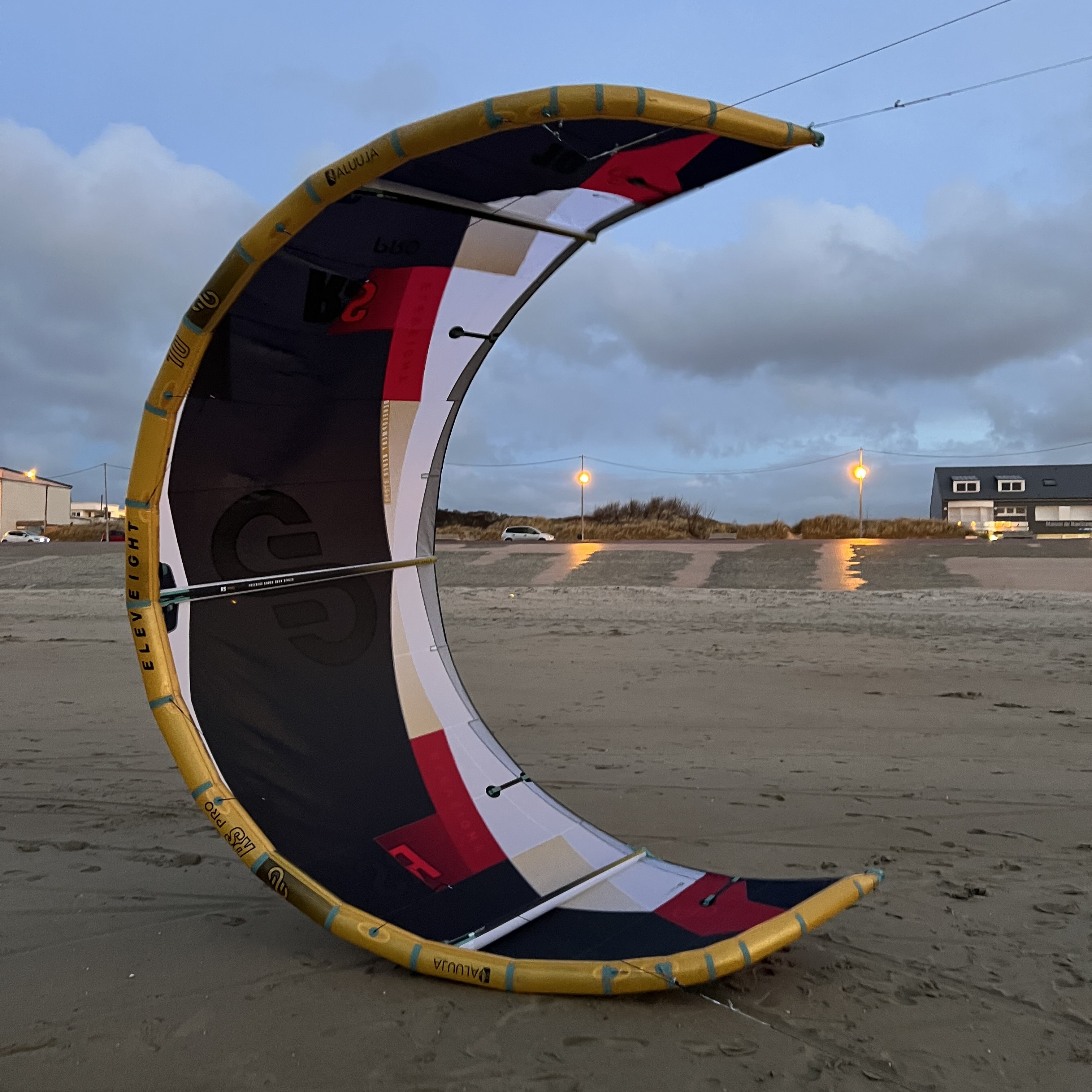 Aile de kitesurf Eleveight RS PRO V1 2025 posée verticalement sur le sable au coucher du soleil, avec maisons et lumières urbaines en arrière-plan – idéale pour le freeride et le big air.