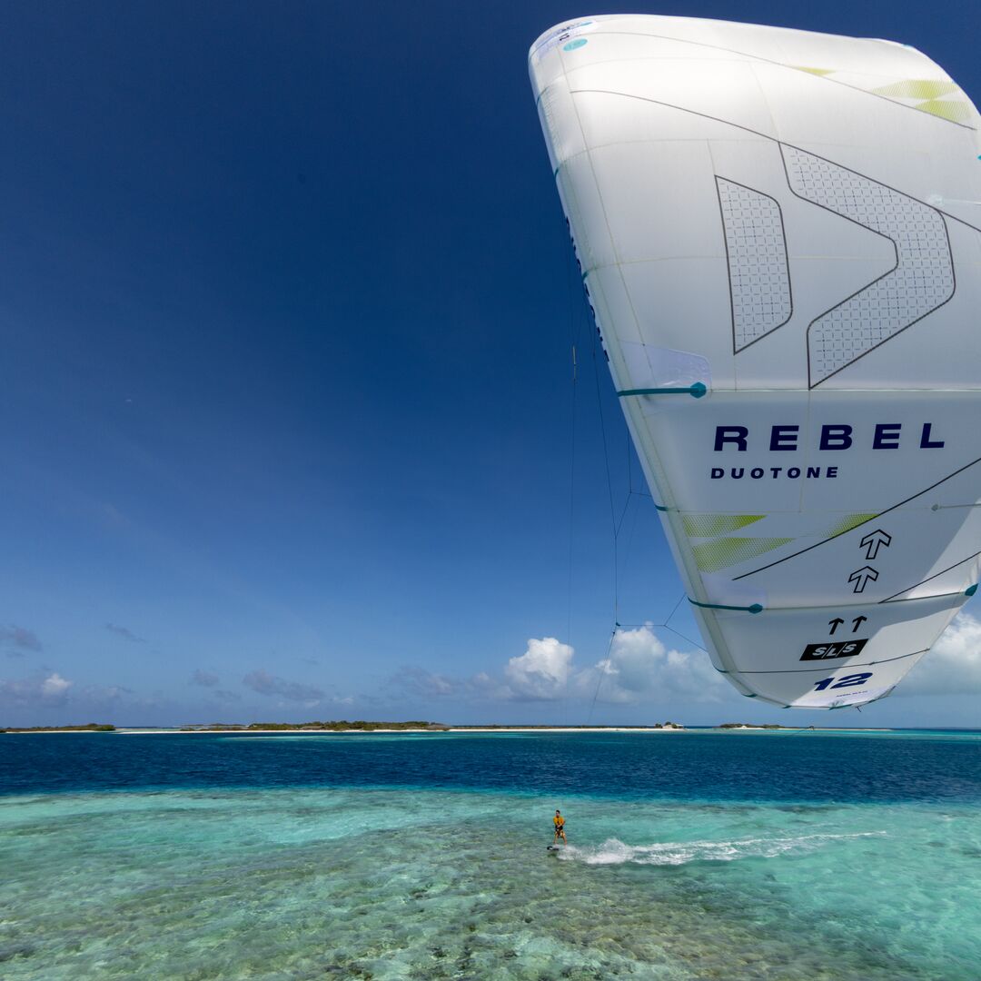 Sur une mer calme et peu profonde, un rider fait du kitesurf avec l'aile Rebel SLS Concept Blue, tandis qu'une plage se dessine au loin sous un ciel ensoleillé.