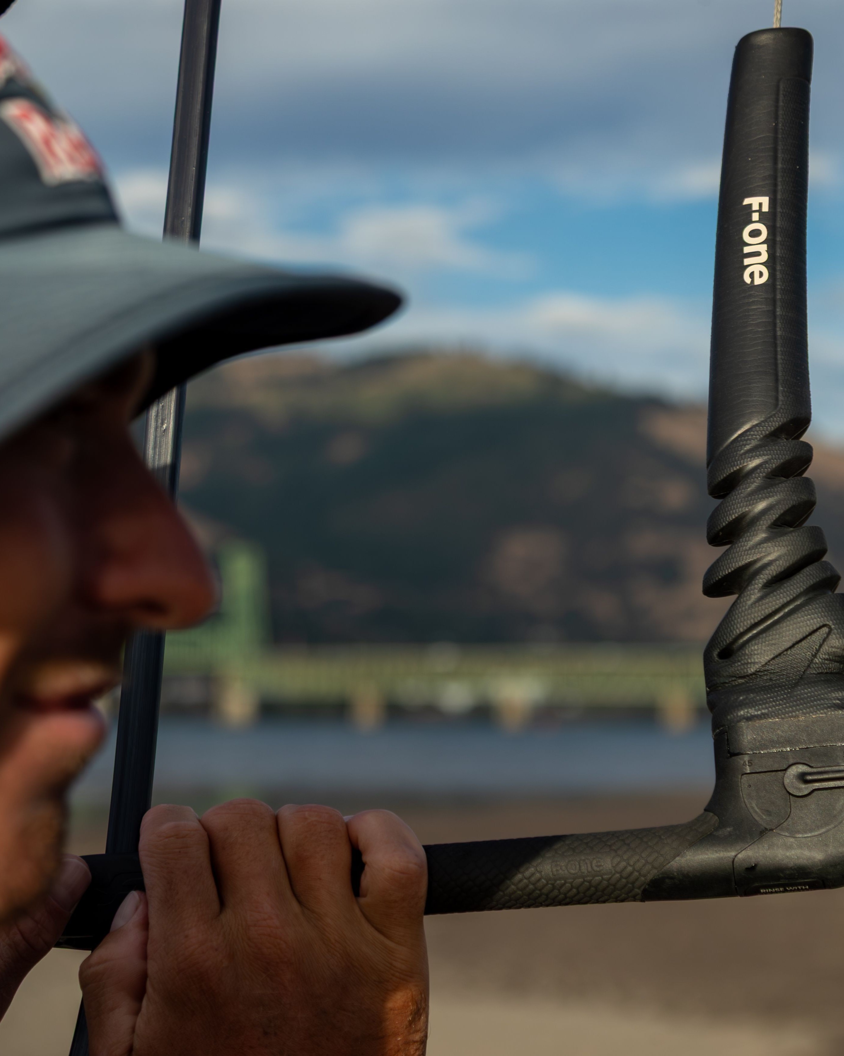 CLOSE-UP du kitesurfer ajustant la barre de kitesurf F-One, avec vue sur les fils et le paysage montagneux en arrière-plan. Idéal pour les passionnés de kitesurf et d'équipements de qualité.